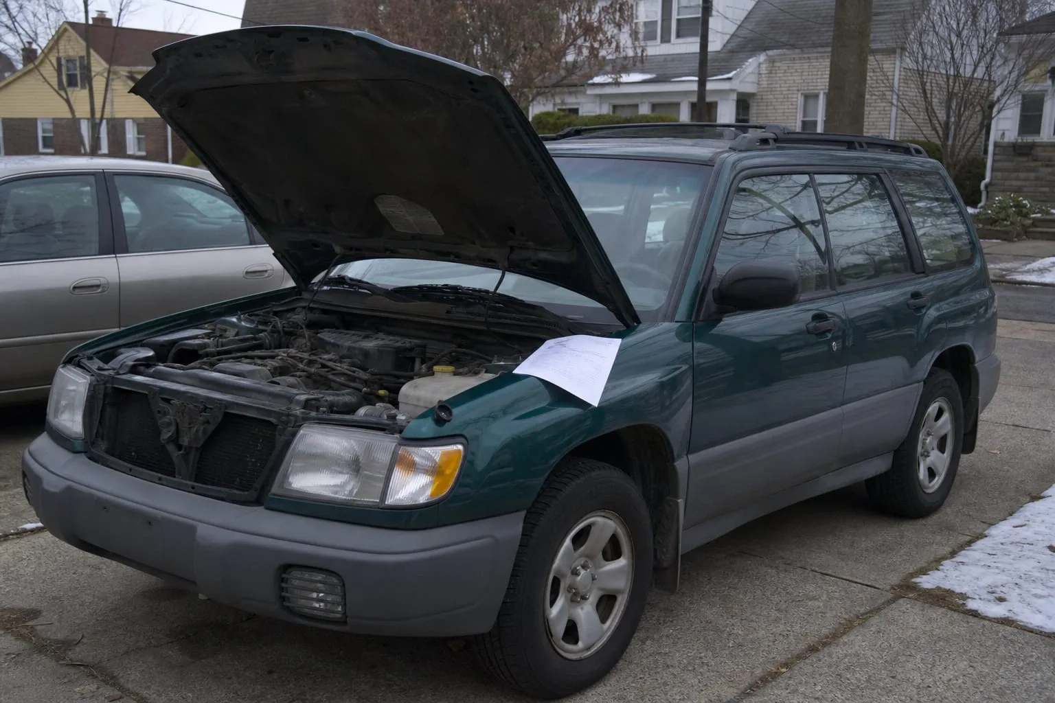 An older compact SUV parked in a suburban driveway on a cold morning, hood raised, coolant reservoir cap off, a printed repair estimate resting on the fender