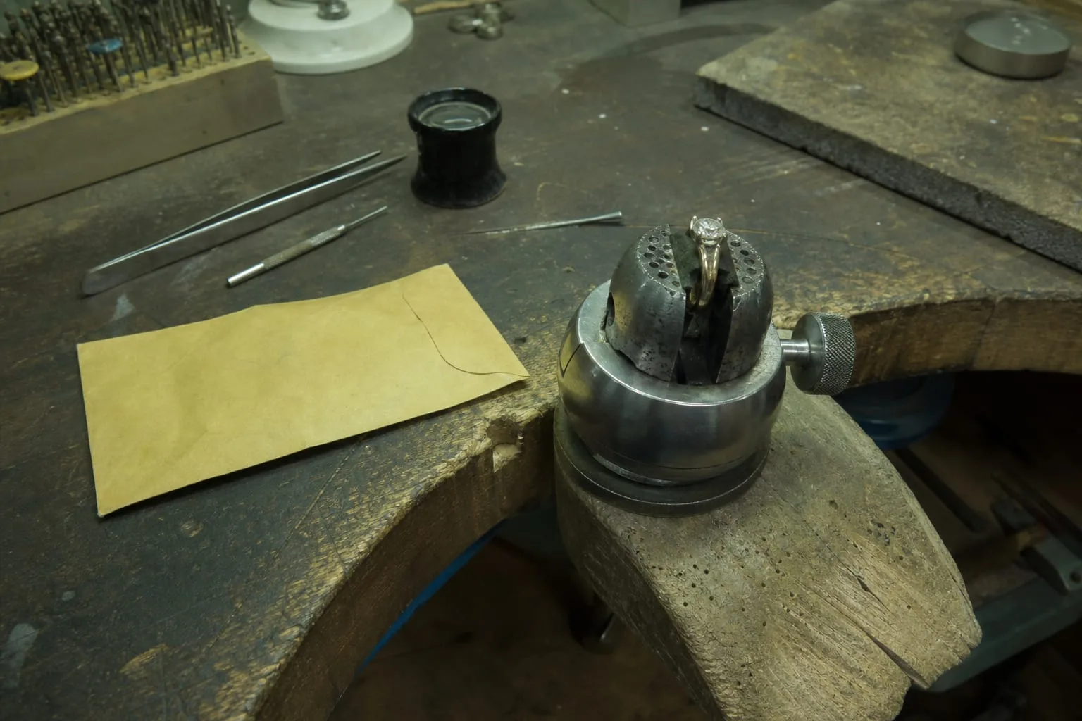 A jeweler's bench with a ring clamped in a setting holder beside a paper repair envelope, small hand tools and a loupe on the worn wooden surface