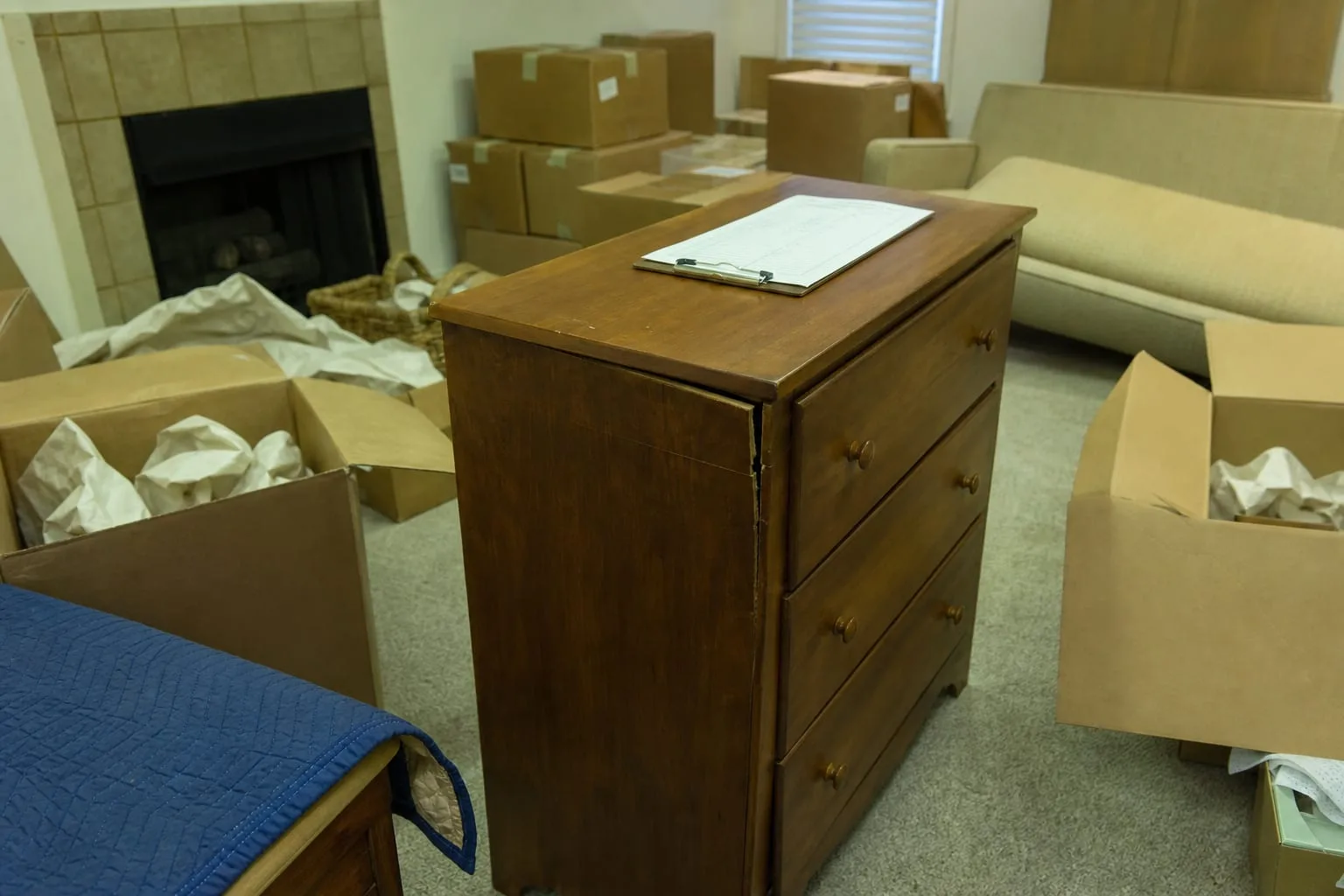 A wooden dresser with a visibly split side rail sitting in a half-unpacked living room among labeled cardboard cartons and packing paper, a clipboard with an...