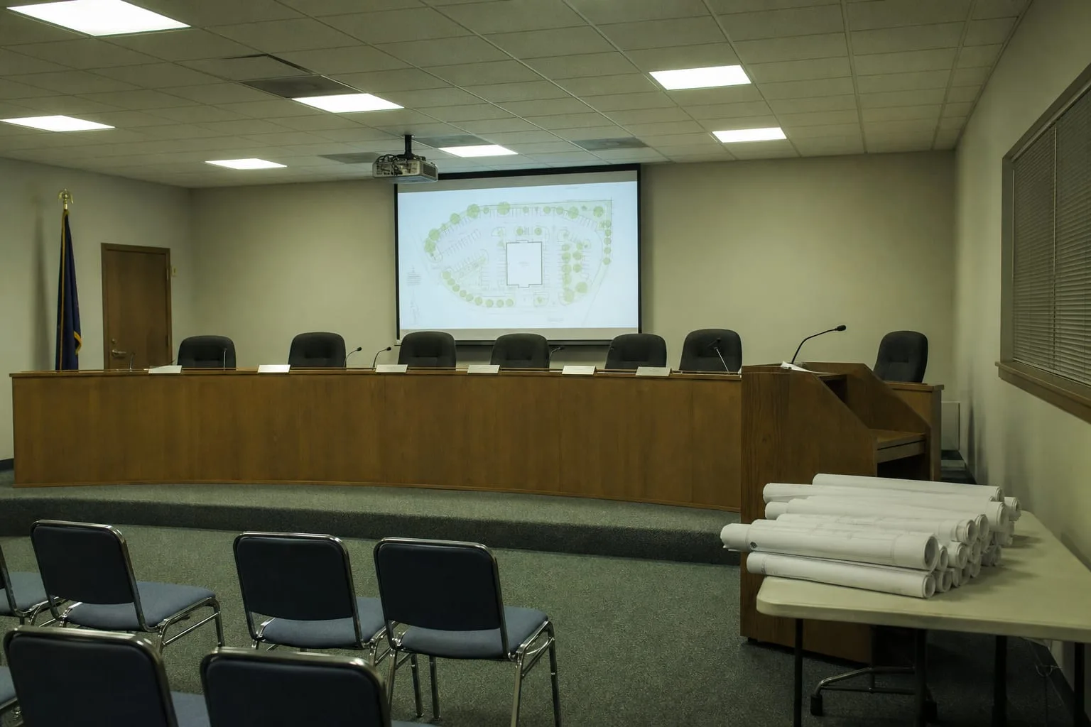 A municipal zoning board hearing room after hours, with a long dais, name placards, a projector screen showing a site plan, and a stack of rolled drawings on...