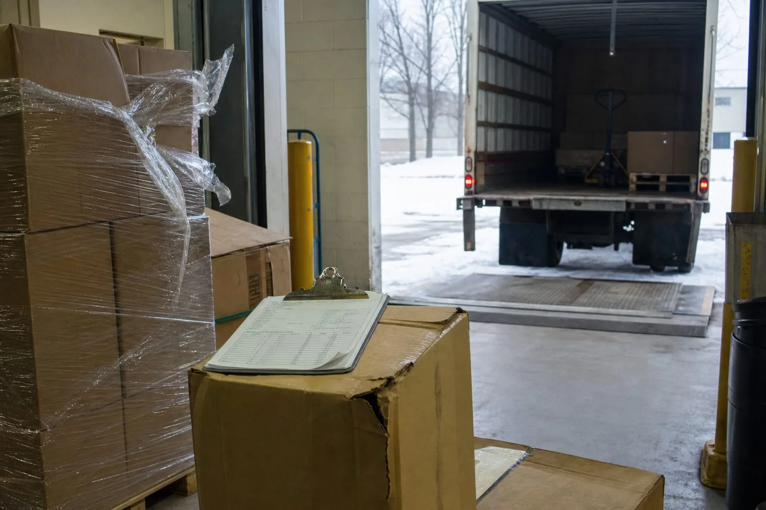 A warehouse receiving dock in winter with a shrink-wrapped pallet partially unwrapped, a clipboard with a delivery receipt resting on a crushed corner of a c...
