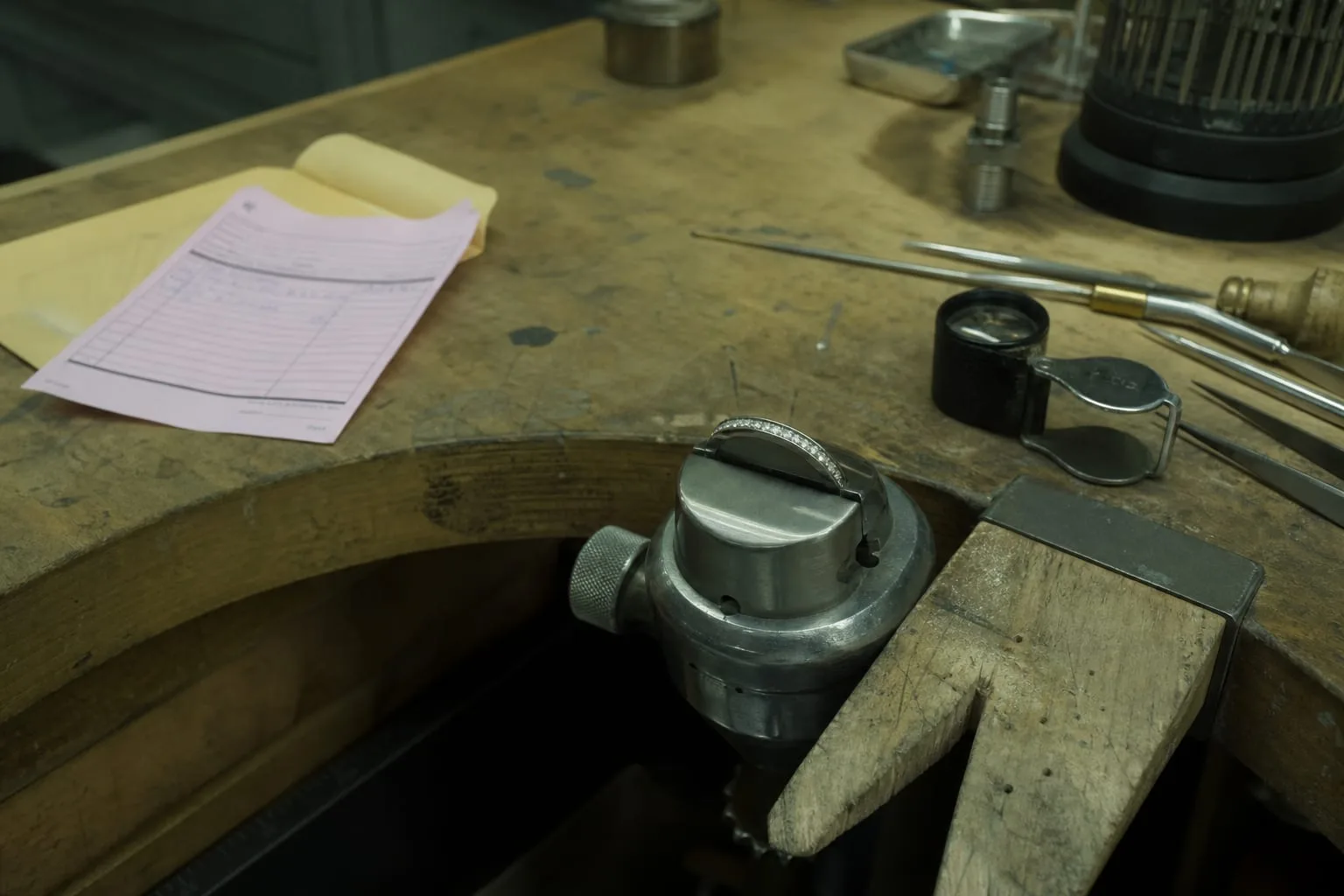 A jeweler's bench with a channel-set diamond band held in a ring clamp, small hand tools and a loupe beside it, a take-in ticket and envelope nearby
